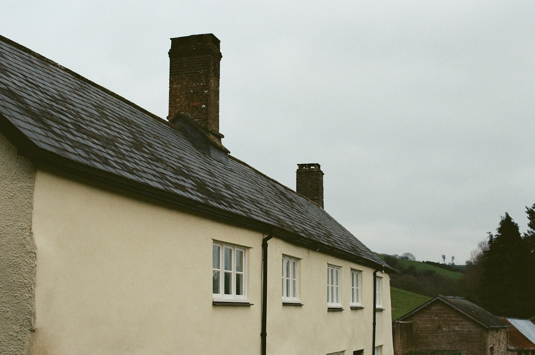Long Devon farmhouse with grey slate roof and brick chimneys under overcast sky